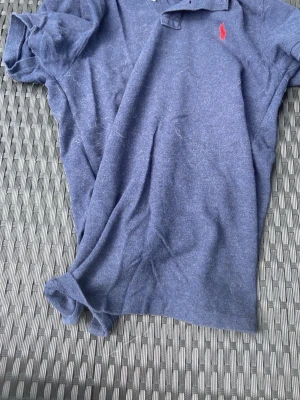 Blå pikétröja från Polo Ralph Lauren - Mörkblå pikétröja från Polo Ralph Lauren med röd broderad logga på bröstet. Slim fit-modell med korta ärmar och klassisk krage med knappar. Tillverkad i mjuk bomull som känns skön mot huden. 