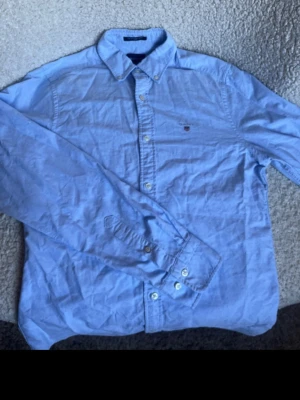 Gant klassisk oxfordskjorta – Ljusblå – Strl S (Slim Fit) - Säljer en klassisk och tidlös ljusblå oxfordskjorta från Gant i passformen "Slim Fit". Ett perfekt basplagg i garderoben som aldrig går ur tiden.  Storlek: S (Slim Fit)  Material: 100% oxfordbomull (kraftigt, slitstarkt och skönt tyg)  Skick: Mycket gott skick, välvårdad och fräsch.  Detaljer: Knappar i kragen (button-down) och den klassiska Gant-skölden broderad på bröstet. Passar lika bra till jeans, chinos eller under en tröja.