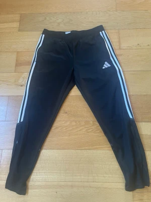 Svarta träningsbyxor från Adidas - Säljer ett par svarta träningsbyxor från Adidas med klassiska vita ränder längs sidorna och Adidas-logga på låret. Väldigt bra skick