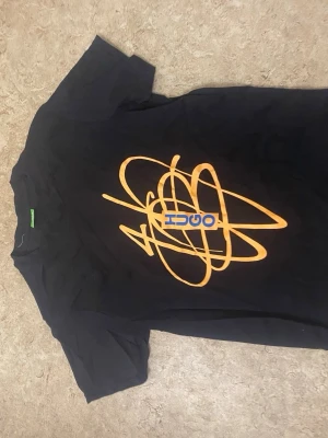 Svart t-shirt med orange tryck - Cool svart t-shirt med ett stort orange grafiskt tryck och blå text på framsidan. Klassisk passform och rund hals. Perfekt för dig som vill sticka ut med en unik design.