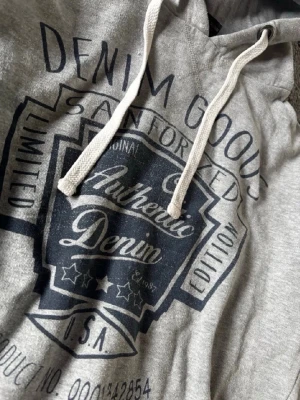 Grå hoodie Denim Goods - Snygg grå hoodie från Denim Goods med stor svart tryck framtill där det står 'Authentic Denim'. Hoodien har huva med vita dragsnören och ribbade muddar vid ärmar och nederkant. Perfekt för en avslappnad streetwear-look.