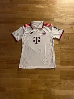 Vit Bayern München Adidas tröja - Säljer en vit Bayern München matchtröja från Adidas med röda detaljer på axlar och ärmslut. Tröjan har klubbmärke, Adidas-logga och Telekoms sponsortryck på bröstet. Klassisk pikékrage och tillverkad i lätt funktionsmaterial.            Fraktar dagen eller samma dag som du köpt. 