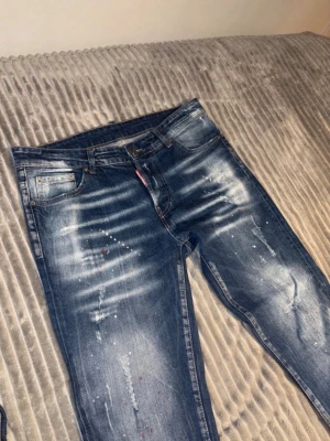 Blå slitna jeans med färgstänk - Säljer ett par blå jeans med coola slitningar och färgstänk på framsidan. Jeansen har klassisk femficksdesign, normal midja och smal passform. Perfekta för dig som gillar en trendig och avslappnad streetstil.