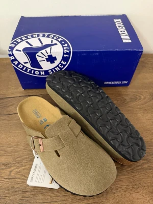Birkenstock Boston tofflor Taupe 38 - Säljer ett par Birkenstock Boston tofflor i mocka med justerbart spänne i metall. Klassisk slip-in modell med mjuk fotbädd och svart räfflad sula. Perfekta för dig som gillar stilren och bekväm design.