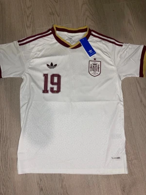 Vit Spanien fotbollströja Adidas - Säljer en vit Spanien fotbollströja från Adidas med vinröda detaljer, nummer 19 på bröstet och Spaniens landslagsemblem. Tröjan har gul och vinröd krage, samt Adidas klassiska ränder på axlarna. Tillverkad i lätt och ventilerande material.