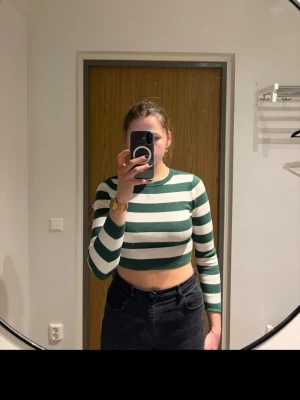 Grönvit randig croppad långärmad topp - Säljer en croppad långärmad topp med breda gröna och vita ränder. Toppen har en rund halsringning och sitter tight mot kroppen. Perfekt för dig som gillar en sportig och trendig stil.