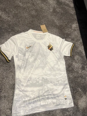 Vit AIK matchtröja från Nike - Säljer en vit AIK matchtröja från Nike med gulddetaljer och klubbmärke på bröstet. Tröjan har ett unikt mönster med Stockholmsmotiv och svarta och gula detaljer vid ärmsluten. Perfekt för dig som vill stötta AIK på match eller träning.