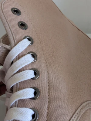 Beige höga tygsneakers - Snygga beige sneakers i canvas med högt skaft och vita snören. Skorna har metallfärgade öljetter och en vit sula som ger en clean look. Perfekta för dig som gillar enkel och stilren design. Passar till det mesta och är lätta att matcha.