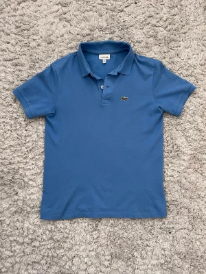 Blå pikétröja från Lacoste - Klassisk blå pikétröja från Lacoste. Perfekt piké inför sommaren med den otroligt snygga blåa färgen. Köpt helt ny och är i nästan helt nytt skick! Storlek 14-15 år (xxs- mindre xs).                  Nypris: Ca 1350 kr. Nu: 399kr!!🤩