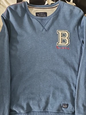 Blå sweatshirt Bondelid med B-logga - Snygg blå sweatshirt från Bondelid med stor vit B-applikation och röd brodyr på bröstet. Tröjan har rund halsringning, ribbade muddar och en liten Bondelid-label vid nederkanten. Perfekt för en avslappnad och stilren look.