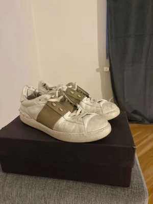 Valentino open  - Snygga vita sneakers från Valentino med bred beige rem över vristen och vita skosnören. Skorna har en klassisk låg siluett och är tillverkade i läder. Perfekta för dig som vill ha en stilren och exklusiv look.