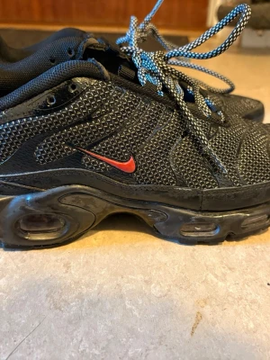 Nike Air Max Plus svarta sneakers - Säljer ett par Nike Air Max Plus sneakers i svart mesh med reflekterande detaljer och röd Swoosh på sidan. Skorna har synlig Air-dämpning i sulan och svarta samt vita mönstrade skosnören. Perfekta för dig som gillar streetwear och sportig stil.