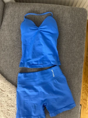 Blått träningsset från DFYNE - Säljer ett blått träningsset från DFYNE med ett linne med tunna axelband och matchande shorts. Setet är gjort i ett stretchigt och mjukt material som passar perfekt för gym eller yoga. Linnet har en lätt rynkning vid bysten och shortsen har bred resår i midjan.