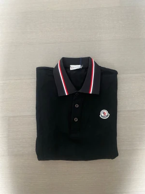 Svart Moncler pikétröja med röd/vit krage - Snygg svart pikétröja från Moncler med klassisk krage som har röda och vita ränder. Tröjan har knappar framtill och Moncler-logga broderad på bröstet. Perfekt för dig som vill ha en stilren och sportig look.