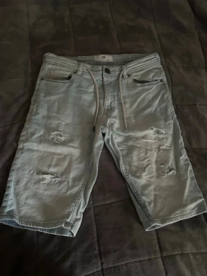 Ljusblå slitna jeansshorts med snörning - Säljer ett par ljusblå jeansshorts från FSBN med slitna detaljer på framsidan och snörning i midjan. Klassisk femficksmodell och normal passform. Perfekta för en avslappnad stil under varmare dagar.