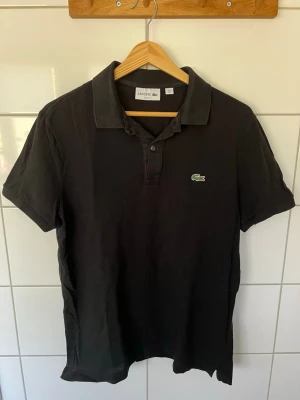 Svart pikétröja från Lacoste - Klassisk svart pikétröja från Lacoste med korta ärmar och krage. Tröjan har knappar framtill och den ikoniska gröna krokodilloggan på bröstet. Tillverkad i mjuk bomull för en bekväm känsla. Storlek L i barnstorlek.