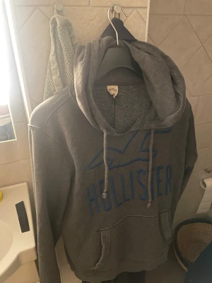 Grå Hollister hoodie med blå logga - Säljer en grå hoodie från Hollister med stor blå logga och fågel på bröstet. Tröjan har huva med snörning och en klassisk magficka. Mjuk och skön bomullsblandning, perfekt för chill dagar. Passar dig som gillar enkelt och stilrent. Lite uppklippt i kragen för mer chill look