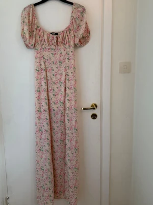 Blommig långklänning från Bik Bok - Romantisk långklänning från Bik Bok i ljusbeige med rosa och grönt blommönster. Klänningen har puffärmar, söt knytning vid bysten och en smickrande v-ringad urringning. Perfekt för sommarens alla tillfällen.