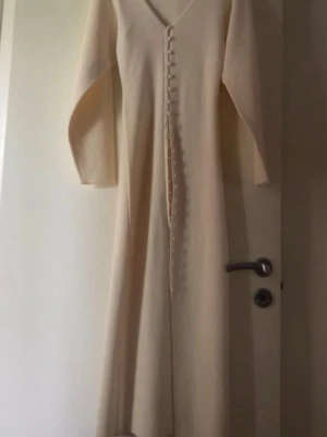 Långärmad beige klänning med knappar - Elegant långklänning i beige med v-ringning och långa ärmar. Klänningen har dekorativa knappar längs hela framsidan och är tillverkad i ett mjukt tyg som ger ett stilrent intryck. Perfekt för dig som gillar minimalistisk och tidlös design. Dock har en knapp gått sönder lite men det medföljer en helt ny knapp :) har aldrig använt den, knappen var sönder när jag beställde den