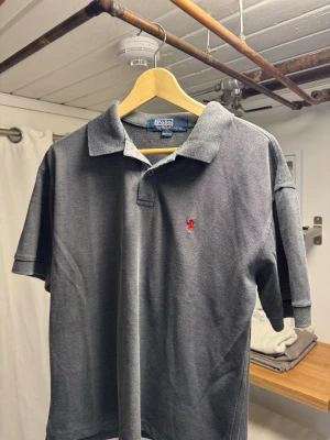 Grå pikétröja från Polo Ralph Lauren - Klassisk grå pikétröja från Polo Ralph Lauren med korta ärmar och röd broderad logga på bröstet. Tillverkad i 100% bomull för en skön och luftig känsla. Perfekt till jeans eller shorts för en avslappnad stil. Små fläckar på ryggen. Storlek L passar M/S