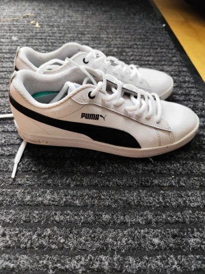 Vita Puma sneakers med svart detalj - Snygga vita sneakers från Puma med klassisk svart sidodetalj och logga. Skorna har lågt skaft, vita snören och ovandel i slätt syntetmaterial. Perfekta för en clean och sportig look.