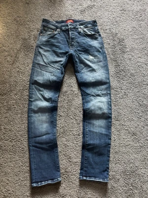 Jack & Jones jeans  - Säljer ett par blå jeans med raka ben och snygga slitningar på låren och knäna. Jeansen har klassisk femficksdesign och kontrastsömmar. Perfekta för en avslappnad och trendig look.
