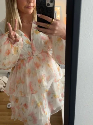 Blommig kortklänning med volanger - Söt kortklänning med volanger och v-ringning i vitt med blommigt mönster i rosa, orange och gult. Klänningen har långa puffärmar och ett luftigt, lager-på-lager tyg som ger en romantisk känsla. Perfekt för sommarens alla tillfällen.