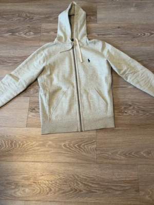 Grå hoodie från Polo Ralph Lauren - Ljusgrå hoodie med dragkedja från Polo Ralph Lauren. Klassisk design med huva och vita dragsnören, två fickor framtill och den ikoniska mörkblå loggan broderad på bröstet. Tillverkad i mjukt bomullsmaterial som känns skönt mot huden.