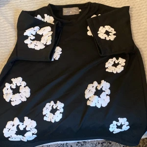 Svart t-shirt med vita blommor från Denim Tears - Säljer en svart t-shirt från Denim Tears med stora vita blommor tryckta över hela tröjan. T-shirten har en klassisk passform och är perfekt för dig som vill sticka ut med ett unikt mönster. Äkta