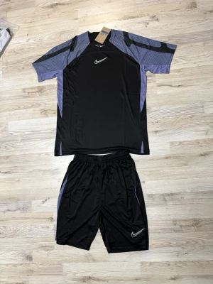 Nike svart träningsset herr - Säljer ett svart träningsset från Nike med t-shirt och shorts. T-shirten har gråblå detaljer på ärmarna och sidorna samt en vit Nike-logga på bröstet. Shortsen har elastisk midja, gråblå detaljer på sidorna och vit Nike-logga. Perfekt för träning eller sport.