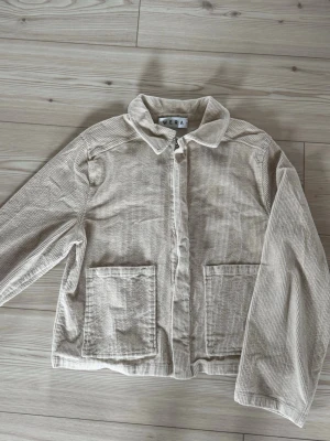 Beige overshirt i manchester från Wera - Säljer en beige overshirt från Wera i mjukt manchestertyg. Jackan har två stora fickor framtill, krage och dragkedja. Perfekt för lager-på-lager och ger en avslappnad look. Materialet är bomull och strukturen är ribbad.