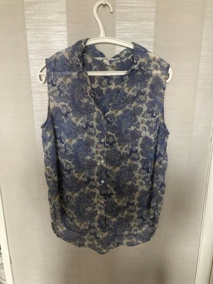 Blommig ärmlös blus från H&M - Snygg ärmlös blus från H&M i storlek 42. Blusen har ett blått och vitt paisleymönster, knappar framtill och klassisk krage. Perfekt till jeans eller kjol för en trendig look. Materialet känns lätt och luftigt.