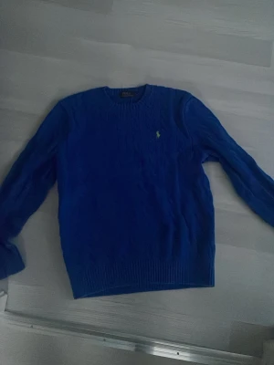 Blå kabelstickad tröja Polo Ralph Lauren - Snygg blå kabelstickad tröja från Polo Ralph Lauren med rund halsringning och klassisk gul broderad logga på bröstet. Tröjan har ribbade muddar vid ärmslut och nederkant. Perfekt för dig som gillar stilren och tidlös design.