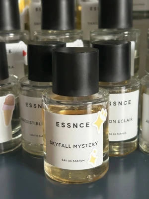 ESSNCE Skyfall Mystery parfym - Skyfall Mystery från ESSNCE är en Eau de Parfum i en stilren, rund glasflaska med svart lock. Etiketten är vit med minimalistisk text och dekorerad med små klistermärken. Doften ger en exklusiv känsla och flaskan har en modern, enkel design.