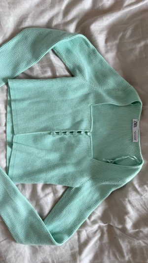 Mintgrön ribbad kofta från Zara - Snygg mintgrön ribbad kofta från Zara med långa ärmar och dekorativa knappar framtill. Koftan har en croppad passform och är tillverkad i ett mjukt, stretchigt material som sitter skönt på kroppen. Perfekt att styla till jeans eller kjol. Det är storlek M men sitter som en XS-S!