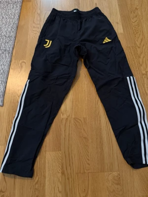 Svarta Juventus träningsbyxor Adidas - Snygga svarta träningsbyxor från Adidas med Juventus-logga i gult och klassiska vita ränder längs benen. Byxorna har resår i midjan och är tillverkade i ett lätt och bekvämt material, perfekta för träning eller chill. Adidas-logga i gult på ena sidan.