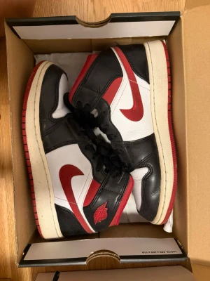Air Jordan 1 Retro mid  OG svart/röd/vit - Säljer ett par Air Jordan 1 Retro mid OG sneakers i svart, rött och vitt läder. Klassisk high-top siluett med röda Swoosh-loggor och vingar på sidan. Tjock vit sula med röd yttersula och svarta skosnören. Perfekta för dig som gillar ikoniska sneakers.