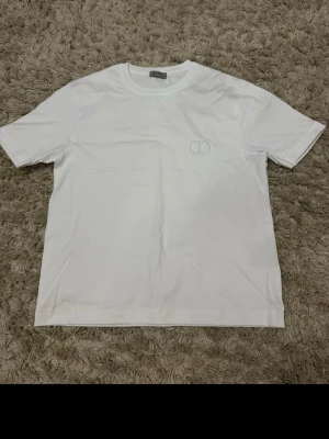 Vit t-shirt från Dior med diskret logga - Säljer en stilren vit t-shirt från Dior med en liten broderad CD-logga på bröstet. T-shirten har klassisk passform, rund halsringning och är tillverkad i mjuk bomull. Perfekt för en clean och trendig look.