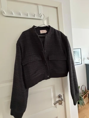 Svart cardiganjacka från Nelly - Stilren svart cardiganjacka från Nelly med strukturerat tyg och två stora bröstfickor. Jackan har rund halsringning, knäppning framtill och ribbade muddar vid ärmslut och nederkant. Perfekt för lager på lager och enkel att matcha.