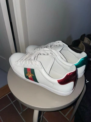 Gucci ace - Säljer ett par vita Gucci sneakers i läder med klassiska gröna och röda ränder på sidan och en guldfärgad bi. Baksidan har metallicröd detalj på ena skon och metallicgrön på den andra, båda med Gucci-logga. Snygg och ikonisk design som sticker ut. Skorna är endast använda 2 gånger så de är i nyskick 