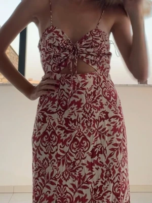 Rödvit sommarklänning med cut-out - Säljer en lång klänning med rödvit bladmönstrad design. Klänningen har tunna axelband, knytning framtill och en snygg cut-out under bysten. Materialet ser ut att vara lätt och luftigt, perfekt för varma dagar. 