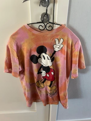 Mickey Mouse t-shirt i tie dye - Färgglad t-shirt från Disney med stort tryck av Mickey Mouse på framsidan. T-shirten har en tie dye-effekt i rosa, orange och gul, samt oversized passform och korta ärmar. Perfekt för dig som gillar retro och tecknade motiv.  