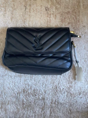 Svart axelväska  - Snygg svart axelväska i quiltat läder med YSL-logga framtill. Väskan har guldfärgade metalldetaljer och justerbar axelrem som medföljer. Perfekt storlek för det viktigaste och ger en lyxig vibe till din outfit.