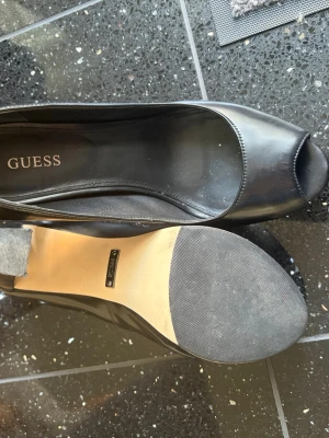 Svarta peep toe pumps från Guess - Snygga svarta pumps från Guess med peep toe och hög platåklack. Skorna är i blankt skinnliknande material och har en diskret logga baktill på hälen. Perfekta för dig som vill ha extra höjd och stilren look.