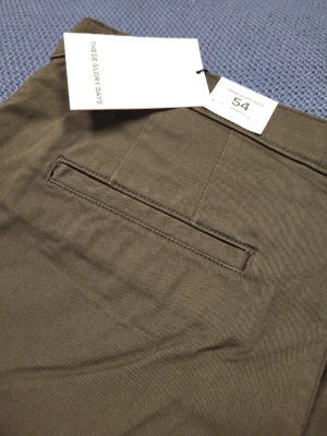 Olivgröna chinos från These Glory Days 54 L - Snygga olivgröna chinos från These Glory Days med klassisk rak passform. Byxorna har bälteshällor, knappgylf och diskreta bakfickor. Tillverkade i ett mjukt och slitstarkt bomullstyg som känns bekvämt hela dagen.