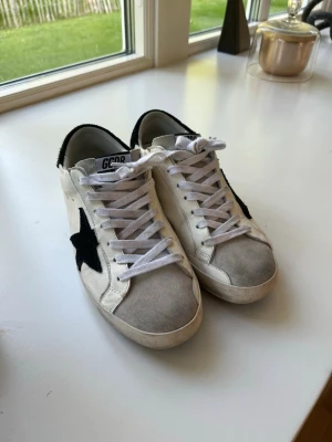 Vita sneakers från Golden Goose - Snygga vita sneakers från Golden Goose med svart stjärna på sidan och svart häl. Skorna har vita skosnören och grå mockadetaljer på tån. Klassisk låg modell med coola kontrastdetaljer och retrovibe. Perfekta för dig som gillar streetstyle och vill sticka ut.