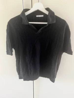 Svart kortärmad bomulltröja  från Zara - Svart tröja  från Zara med korta ärmar och klassisk krage. Tröjan  har en lätt ribbad struktur och är tillverkad i ett mjukt material som känns skönt mot huden. Perfekt för en avslappnad och stilren look.