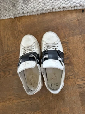 Valentino vita sneakers med svart band - Snygga vita sneakers från Valentino med ett brett svart band över vristen och coola nitar på sulan. Skorna har klassisk låg profil och är tillverkade i läder med vita snören. Skorna lite skitiga men tvättar upp innna köp, köpte ny från merchsweden 7200