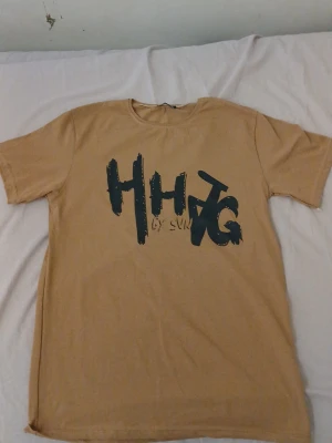 Beige t-shirt med tryck HHJG - Snygg beige t-shirt med stort svart tryck på bröstet där det står HHJG i en cool, grafisk stil. T-shirten har rund hals och korta ärmar. Perfekt för dig som gillar streetwear och vill ha en enkel men trendig look.