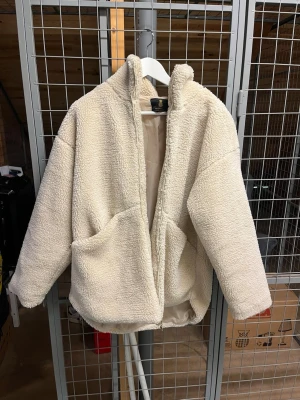 Beige teddyjacka med dragkedja - Mysig beige teddyjacka med hög krage och dragkedja framtill. Jackan har två stora fickor på sidorna och är tillverkad i fluffigt teddy-material som ger en avslappnad och trendig look. Perfekt för att hålla värmen under kyliga dagar.